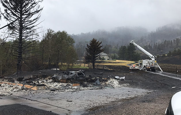 A burned car and tree is in the foreground with a bucket truck in the background